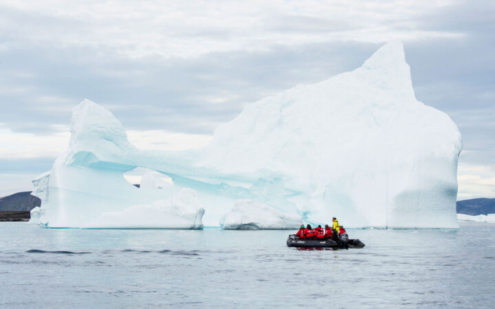 Thule region travelers in red parkas ride a Zodiac boat beside a massive iceberg in calm water under cloudy skies.
