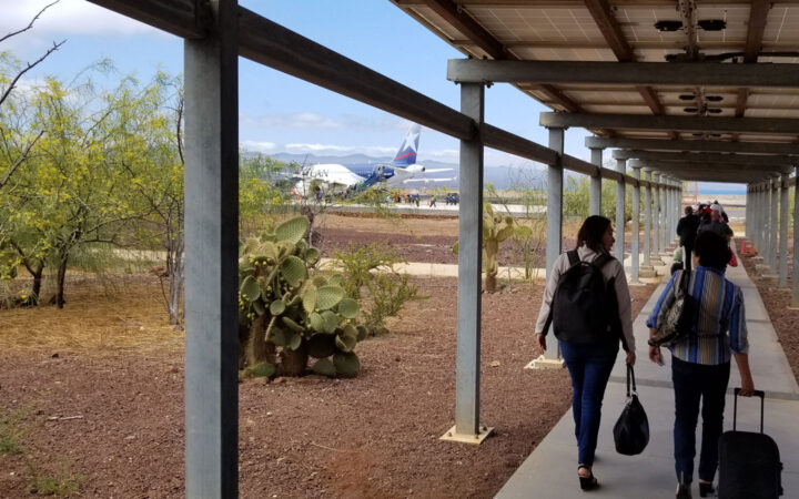 Travelers walk outside the Galapagos airport under an awning walkway toward a plane on Baltra Island.