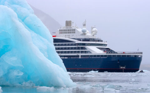 Le Commandant Charcot ship with blue hull & white upper decks peeks around bright blue iceberg on a Greenland to Svalbard cruise.