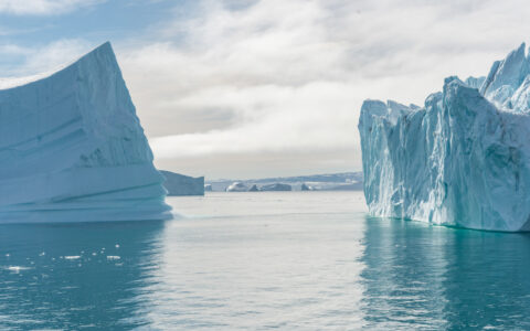 2 large icebergs sit in calm glacial water under cloudy sky, seen on an In the Ice of the Arctic, Greenland to Svalbard cruise.