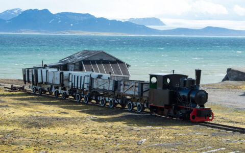 Abandoned train sits on a track among yellow tundra backed by turquoise water with whitecaps & mountains behind, in Svalbard.