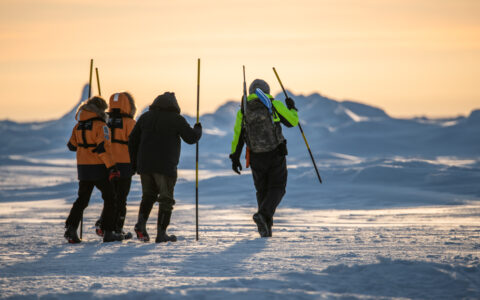 Charcot North Pole travelers in orange coats walk with long sticks over ice & snow, led by guide in neon & black coat at dusk.