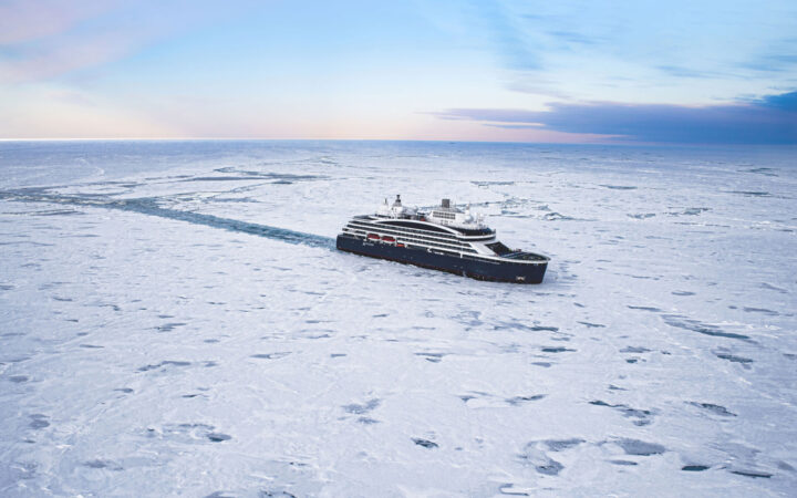 Modern small ship Le Commandant Charcot with dark blue hull & white upper decks crushes through ice at sunset at the North Pole.