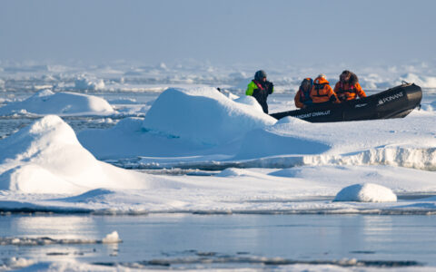 Zodiac boat with North Pole travelers in orange parkas & guide in neon green & black parka sits on an iceberg in the sun.