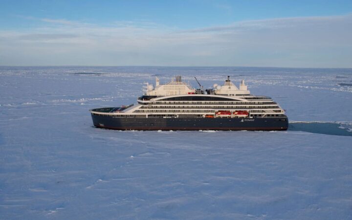 Aerial view of expedition ship with dark blue hull & white upper decks, cruising through ice under a blue sky.