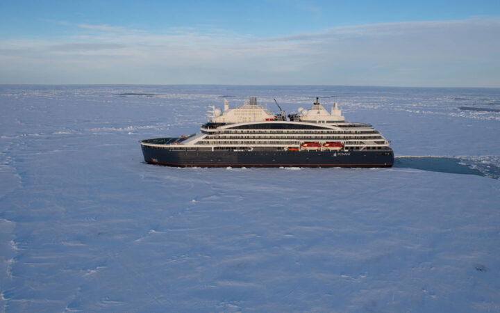 Aerial view of expedition ship with dark blue hull & white upper decks, cruising through ice under a blue sky.