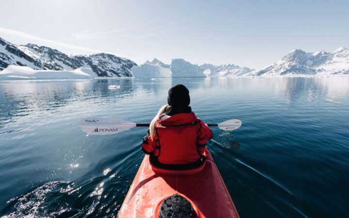 Female in red parka paddles red kayak through calm Arctic ocean surrounded by snow covered jagged mountains in Greenland.