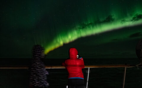 Travelers in red & black jackets stand on deck of a ship & photograph a green aurora borealis at night in East Greenland.
