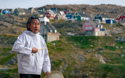 Local Greenlandic man in white anorak stands & beats a hand drum beside a hill with colorful homes in the Arctic.