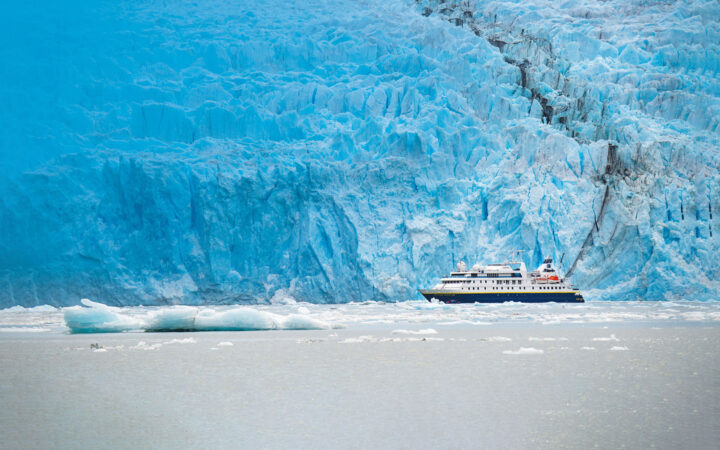 Small blue & white expedition ship on a cruise from Chilean fjords to Antarctica, beside a wall of blue ice.