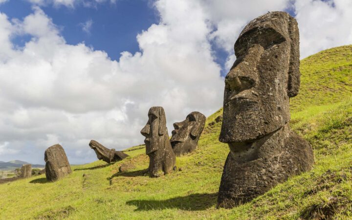 a grassy hillside in easter island chile with several moai statues sitting on it with puffy clouds behind them