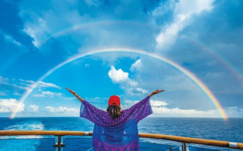 Woman stands at bow of ship & raises both arms toward a double rainbow over the sea on a sunny day near Easter Island.