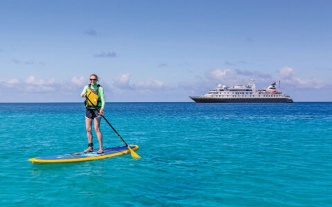 Woman stand-up paddleboarding over turquoise sea with a small ship behind at Tahanea Atoll, Tuamotus, French Polynesia.
