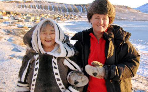 Older woman in warm Inuit garb stands beside younger woman in brown jacket & furry hat on a sunny day in the Thule region.
