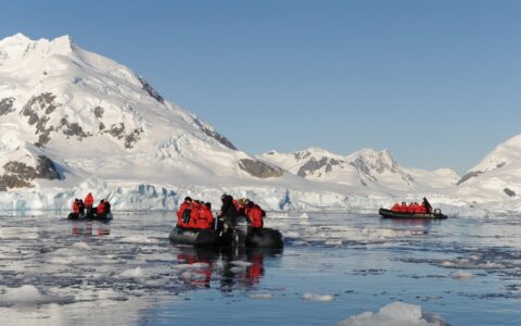 3 black Zodiac boats with polar travelers in red jackets, cruise among ice chunks beside snow-covered hills on a sunny day.