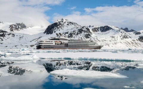 A luxury ship colored white and silver with a red stripe float in ice covered water in front of snowy mountain range in Svalbard