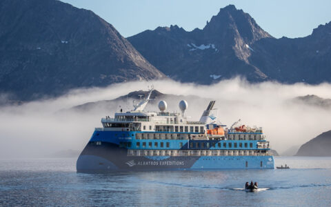 Modern small ship with blue & white exterior sits in foggy fjord under jagged peaks as Zodiac boat cruises away from the ship.