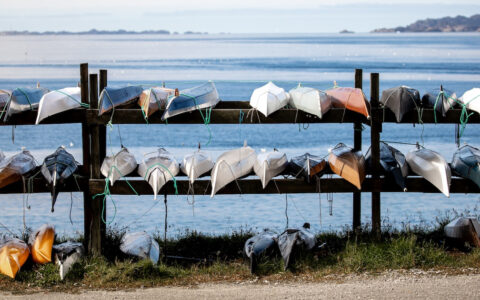 Small modern kayaks lay upside down on wooden storage racks beside blue sea in Greenland, seen on a Four Arctic Islands cruise.