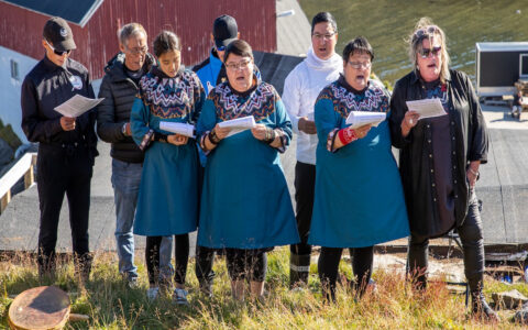 Small group of East Greenland locals in modern garb & traditional blue dresses stands atop a grassy hillside in the sun & sings.