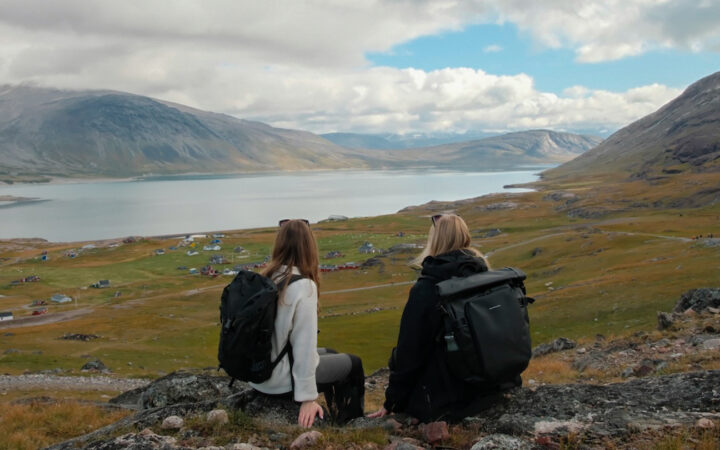2 women with black backpacks sit atop a rocky overlook & view green hills, rounded mountains, colorful homes & calm fjord.