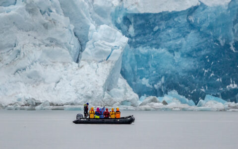 Zodiac with Four Arctic Islands cruise guests motors past a massive blue & white glacier face in East Greenland.