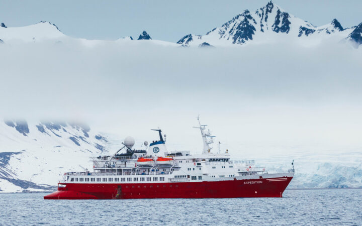 G Expedition ship with red hull & white upper decks sits in calm sea under clouds in snowy Antarctic landscape.