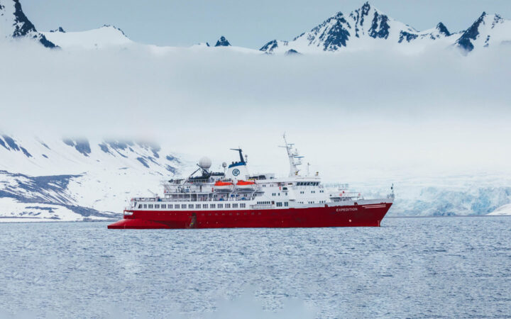G Expedition ship with red hull & white upper decks sits in calm sea under clouds in snowy Antarctic landscape.