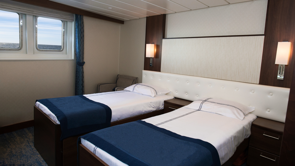 Deluxe Twin cabin on G Expedition ship with 2 twin beds in blue & white linens, padded white headboard & 2 small windows.