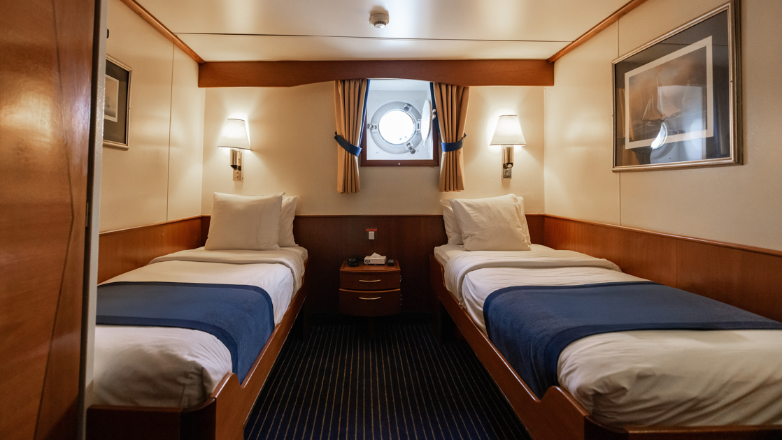 Main Deck Porthole Twin cabin on G Expedition ship with 2 twin beds in blue & white linens, wood accents & 1 porthole.