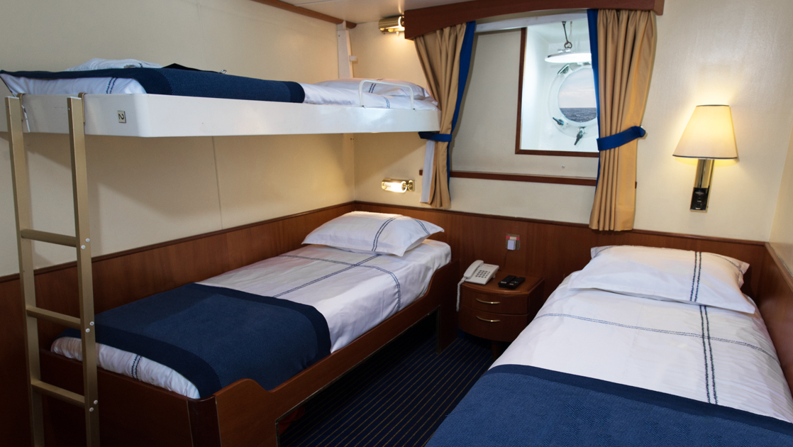 Main Deck Triple cabin on G Expedition ship with bunkbeds & 1 lower twin bed in blue & white linens, wood accents & porthole.