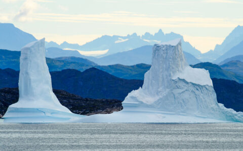 Massive blue & white iceberg growler pieces rise like statues out of the sea, backed by rows of mountains in Greenland.