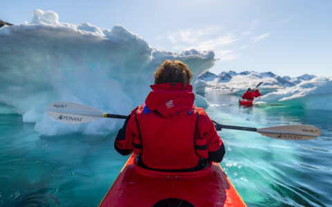 Greenland to Canada cruise traveler in red jacket & life vest paddles a red kayak among iceberg growlers on a sunny day.
