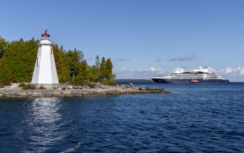 Small blue & white expedition ship sits offshore from white & red lighthouse backed by green forest in eastern Canada.