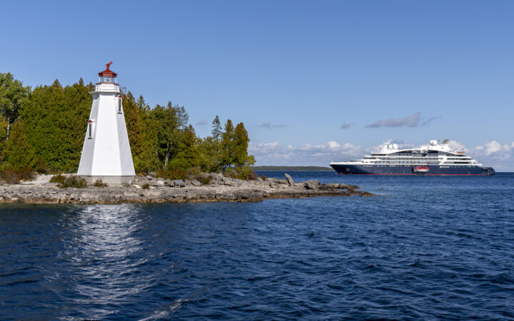 Small blue & white expedition ship sits offshore from white & red lighthouse backed by green forest in eastern Canada.