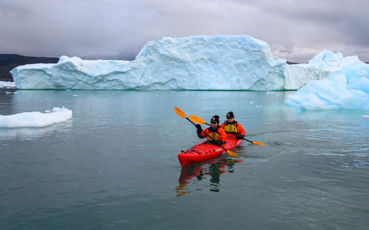 West Greenland travelers paddle a red tandem kayak among large blue iceberg bits under a cloudy sky.