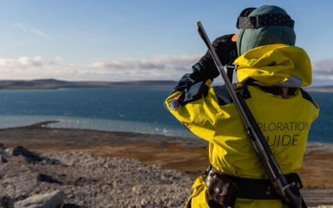 Expedition guide in yellow jacket & hat, gloves & goggles looks through binoculars while wearing a rifle in Nunavut, Canada.