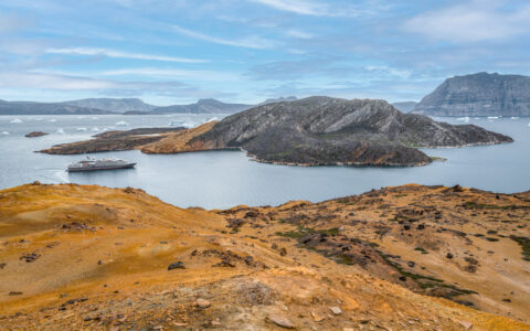 Small silver & white expedition ship sits in glassy fjord between golden islands & others made of steep cliffs in Greenland.