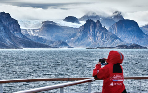 Cruise traveler from Greenland to Canada's East Coast in red jacket stands on deck of ship & views jagged peaks in the distance.