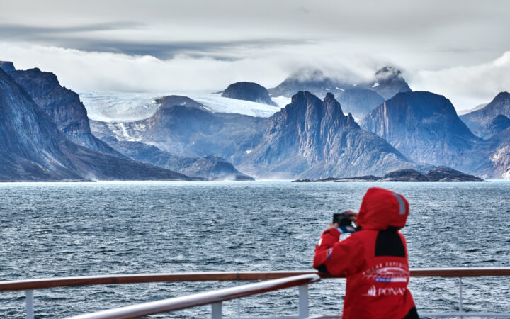 Cruise traveler from Greenland to Canada's East Coast in red jacket stands on deck of ship & views jagged peaks in the distance.