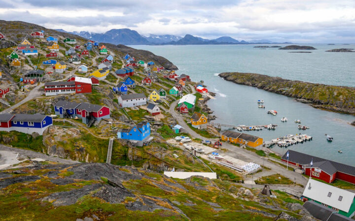 Aerial view of small town with colorful buildings sitting atop a green hillside by small marina under a cloudy sky in Greenland.