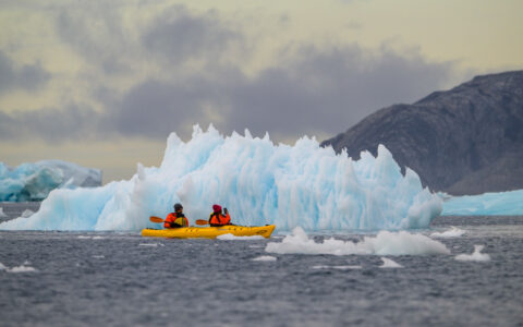Tandem kayakers paddle a yellow boat past a jagged iceberg bit in calm sea on a cloudy day on the Greenland Explorer cruise.