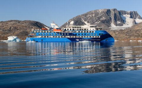Small, modern blue & white ship with pointed bow sits in glassy sea beside snowy stone peaks & green hills in Greenland.