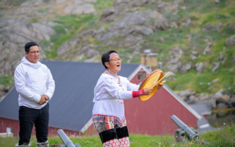 Man stands & watches woman sing & play a round drum in colorful Inuit clothing amid green hills on the Greenland Explorer cruise.