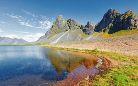 Remote shoreline of green grass & beige sand beneath jagged peaks overlooking glassy water on a sunny day in Iceland.
