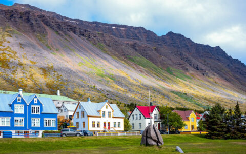 Row of traditional Icelandic homes in blue, white & red fronted by green grass & backed by tall volcanic peaks under clouds.