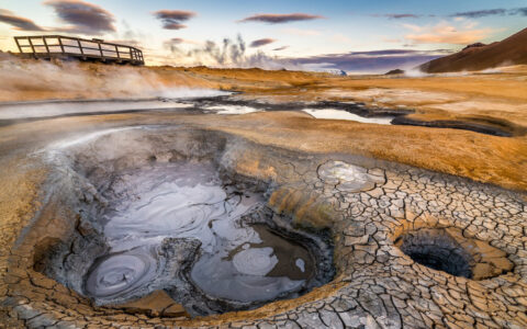 Boiling geothermal pool beside cracked desert landscape & wooden viewing platform, seen at dusk, in Iceland.