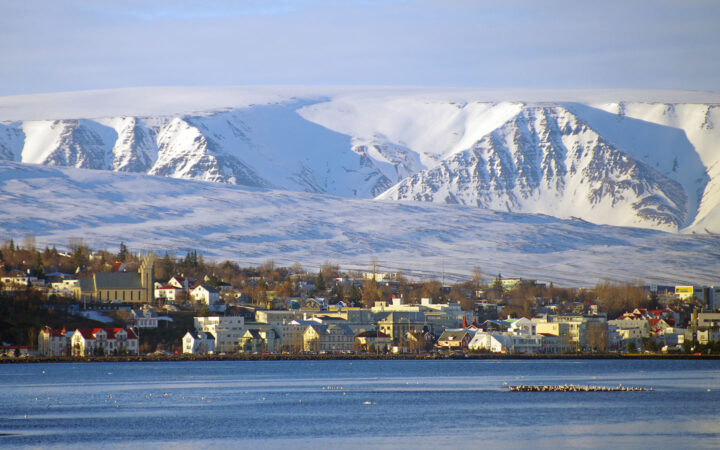 Seaside town of Akureyri, Iceland in winter with beige & white buildings backed by tall, snowy mountains.