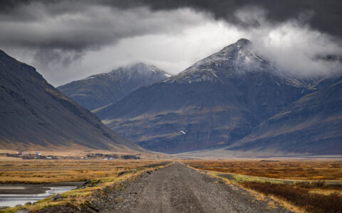 Secluded gravel road in Iceland leads past autumnal flatlands to tall volcanic mountains in the clouds.
