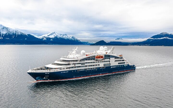 Aerial view of Le Bellot luxury expedition ship with dark blue hull & white upper decks, cruising through calm waters with snow-covered peaks in the background.