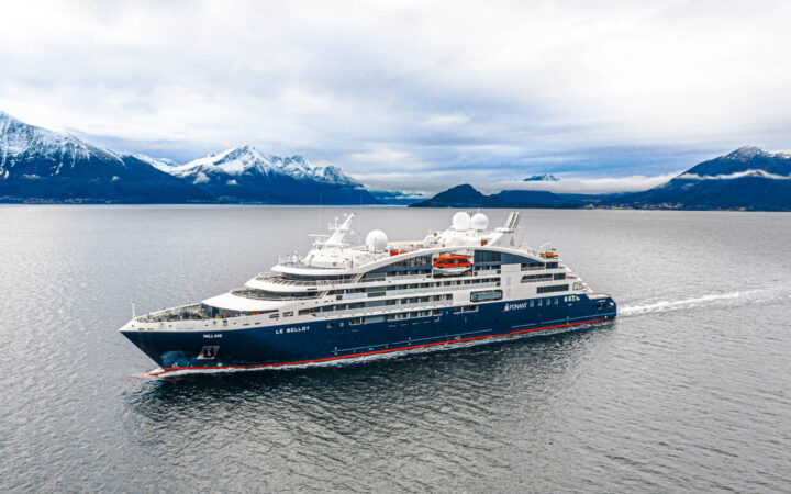 Aerial view of Le Bellot luxury expedition ship with dark blue hull & white upper decks, cruising through calm waters with snow-covered peaks in the background.